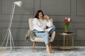 A smiling business woman holding glasses near her mouth, wearing a white shirt and jeans, is sitting on a beige chair with a lamp Royalty Free Stock Photo