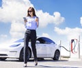 Smiling business woman walking on city parking near electric car, charging automobile battery from small city station, drinking c