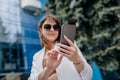 Smiling business woman in white suit and sunglasses using phone during break standing near modern office building Royalty Free Stock Photo