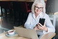 Smiling businesswoman is sitting at table in front of laptop and using smartphone. Pensioner freelancer works. Royalty Free Stock Photo