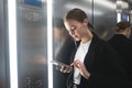 Smiling businesswoman is using her smartphone with headphones in the elevator. Cheerful office worker is texting on the phone and Royalty Free Stock Photo