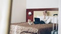 Smiling businesswoman in white shirt using on laptop and talking at mobile phone while lying on bed in hotel room Royalty Free Stock Photo