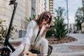 Smiling businesswoman in white suit sitting on electric scooter and records an audio message during walking in city