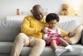 Smiling busy african american senior grandfather and little girl sitting on couch, granddaughter playing in online game Royalty Free Stock Photo