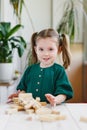 Smiling canny cute child in green dress and broken wooden jenga tower on a table. Indoor activities concept