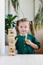 Smiling canny cute child in green dress looking at a wooden jenga tower standing on a table
