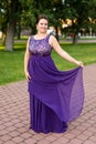 Smiling Caucasian brunette woman is touching hem of purple floor-length dress in park Royalty Free Stock Photo