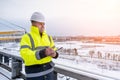 A smiling caucasian builder in a white hard hat and yellow fluorescent jacket holds clipboard