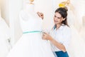 Smiling caucasian designer woman or is bridal shop owner with tape measure measures wedding dress on mannequin in workshop, Royalty Free Stock Photo