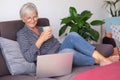 Smiling caucasian elderly woman sitting on sofa at home holding a coffee cup while browsing on laptop Royalty Free Stock Photo