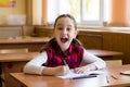 Smiling caucasian girl sitting at desk in class room and happily shouts. Portrait of young pre schoolgirl. Happy pupil