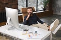 Smiling caucasian man wearing casual wear sitting at the armchair with his legs at the table and using computer at Royalty Free Stock Photo