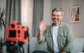 Smiling caucasian mature male with beard waving hand at camera, shooting video in living room interior