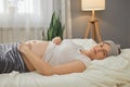 Smiling Caucasian pregnant woman wearing blindfold and pajama lying on bed and touching her bare belly at home looking at camera Royalty Free Stock Photo