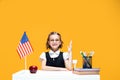 Smiling caucasian schoolgirl raising hand sitting at the desk during lesson. English lesson USA flag