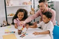 smiling caucasian teacher in eyeglasses and african american kids drawing pictures with paints together Royalty Free Stock Photo