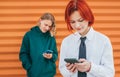 Smiling caucasian teenage girl portrait with red dyed hair and friend boy behind browsing their smartphone devices. Careless young Royalty Free Stock Photo