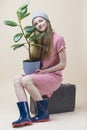 Smiling Caucasian Teenager Girl In Red Checked Dress and Grey Winter Hat Posing With Ficus On Old Suitcase and Rubber Boots Over