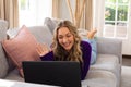 Smiling caucasian woman lying on couch making laptop video call, waving in sunny living room Royalty Free Stock Photo