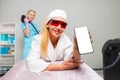 Smiling caucasian woman in protective eyeglasses lies on a couch holding a cellphone with mock up. Cosmetologist does an
