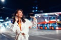 Smiling caucasian woman using mobile phone and earphones while walking in the city at night, free space