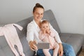 Smiling Caucasian woman wearing white shirt and jeans sitting on sofa with baby daughter, holding mobile phone and taking selfie Royalty Free Stock Photo