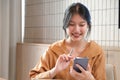 Smiling and charming Asian woman sit at her desk with a smartphone playing social media applications and transacting at her desk Royalty Free Stock Photo