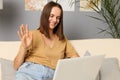 Smiling cheerful brown haired woman wearing beige T-shirt and jeans sitting on couch and working on portable computer in home