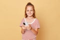 Smiling cheerful cute little girl wearing striped T-shirt standing  over beige background using mobile phone watching Royalty Free Stock Photo