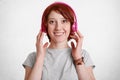 Smiling cheerful gnger femal with freckled face, listens radio or music with modern headphones, smiles broadly, dressed in casual Royalty Free Stock Photo