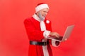 Smiling cheerful man in eyeglasses and santa claus costume working on laptop, sending holidays greeting cards Royalty Free Stock Photo