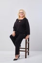 Smiling middle aged woman in black outfit sitting on stool over white background