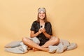 Smiling cheerful positive young girl sitting floor with blanket wearing black pajama and sleep eye mask isolated over beige Royalty Free Stock Photo