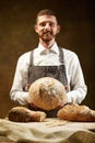 Smiling chef confectioner in apron hold and show warm round homemade bread over studio background. Whole grain rye bread