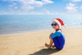 smiling child boy in santa hat, sunglasses and swimsuit sitting on the sand beach Royalty Free Stock Photo