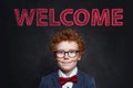 Smiling child with ginger hair portrait. Cute little boy in blue suit against blackboard with welcome inscription