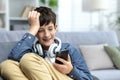 A smiling child of a teenage boy in headphones sitting on the sofa at home, resting and using the phone. Listens to Royalty Free Stock Photo