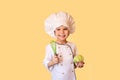 Smiling child in white chef uniform,holding a cabbage and a whisk in his hands. Cheerful yellow background Royalty Free Stock Photo