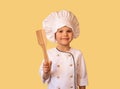 Smiling child in white chef uniform,holding wooden spatula in hands. Cheerful yellow background Royalty Free Stock Photo