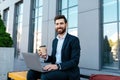 Smiling confident european millennial bearded businessman in suit work on laptop, drink coffee on bench