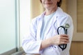 Smiling confident female doctor in white workwear holding stethoscope, standing with arms crossed by window in clinic hospital. Royalty Free Stock Photo