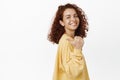 Smiling confident woman with red curly hairstyle, pointing finger behind her, showing smth left, standing over white Royalty Free Stock Photo