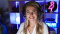 Smiling, confident young blonde streamer, radiantly beautiful at her gaming room, playfully engaged in futuristic cyber gaming