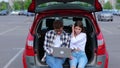 Smiling couple looking at laptop screen while sitting on the trunk car on the parking outdoors. Tansportation