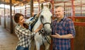 Smiling couple with white horse standing at stabling indoor Royalty Free Stock Photo