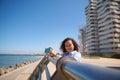 Smiling curly haired multi ethnic pretty woman with mobile phone, standing at seacoast, smiling looking at camera Royalty Free Stock Photo