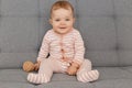 Smiling cute charming baby girl wearing red striped sleeper playing alone at ho me, sitting on gray couch with wooden toy in hands Royalty Free Stock Photo