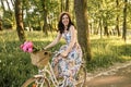 Smiling cute happy girl in dress and hat riding retro bicycle with flowers in the buncket. Green sunny park is around Royalty Free Stock Photo