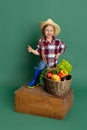 Smiling cute little girl, emotive kid in image of farmer, gardener standing near giant basket of vegetables isolated on