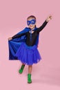 Smiling cute little girl in superhero costume holding his hand and pointing up over pink background